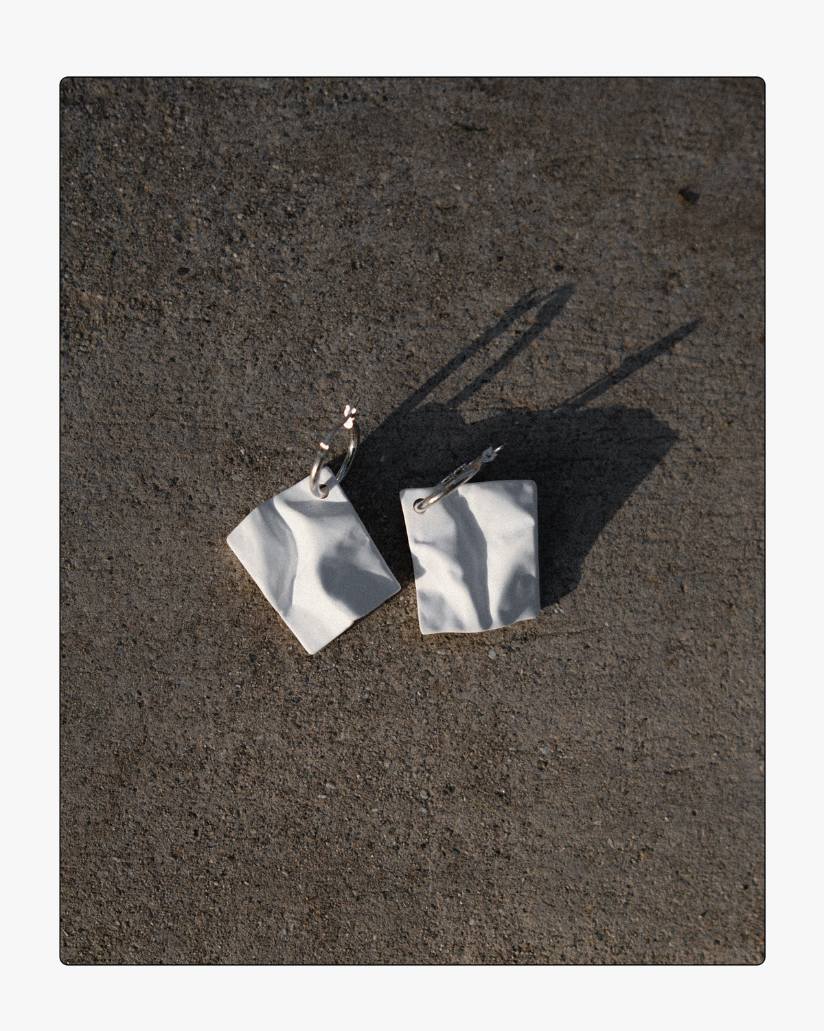 Modern white ceramic paper-texture drop earrings on silver hoops displayed on raw concrete in high-contrast sunlight.