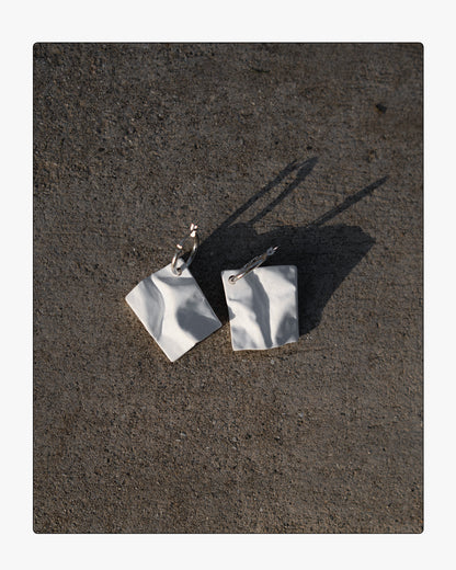 Modern white ceramic paper-texture drop earrings on silver hoops displayed on raw concrete in high-contrast sunlight.