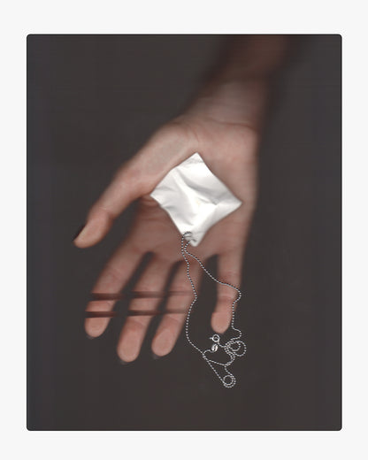 Hand holding a hand-cast white ceramic paper-texture necklace pendant, showing scale and chain attachment against a soft grey background.