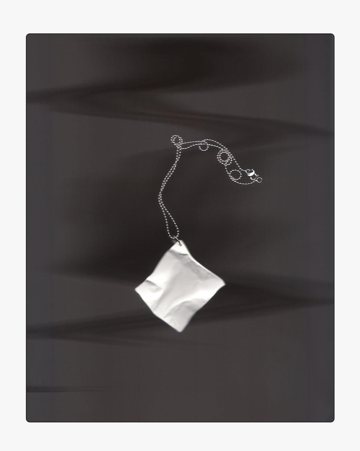 Modern white ceramic paper-texture necklace pendant displayed on a dark surface under dramatic lighting and long shadows.