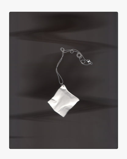 Modern white ceramic paper-texture necklace pendant displayed on a dark surface under dramatic lighting and long shadows.