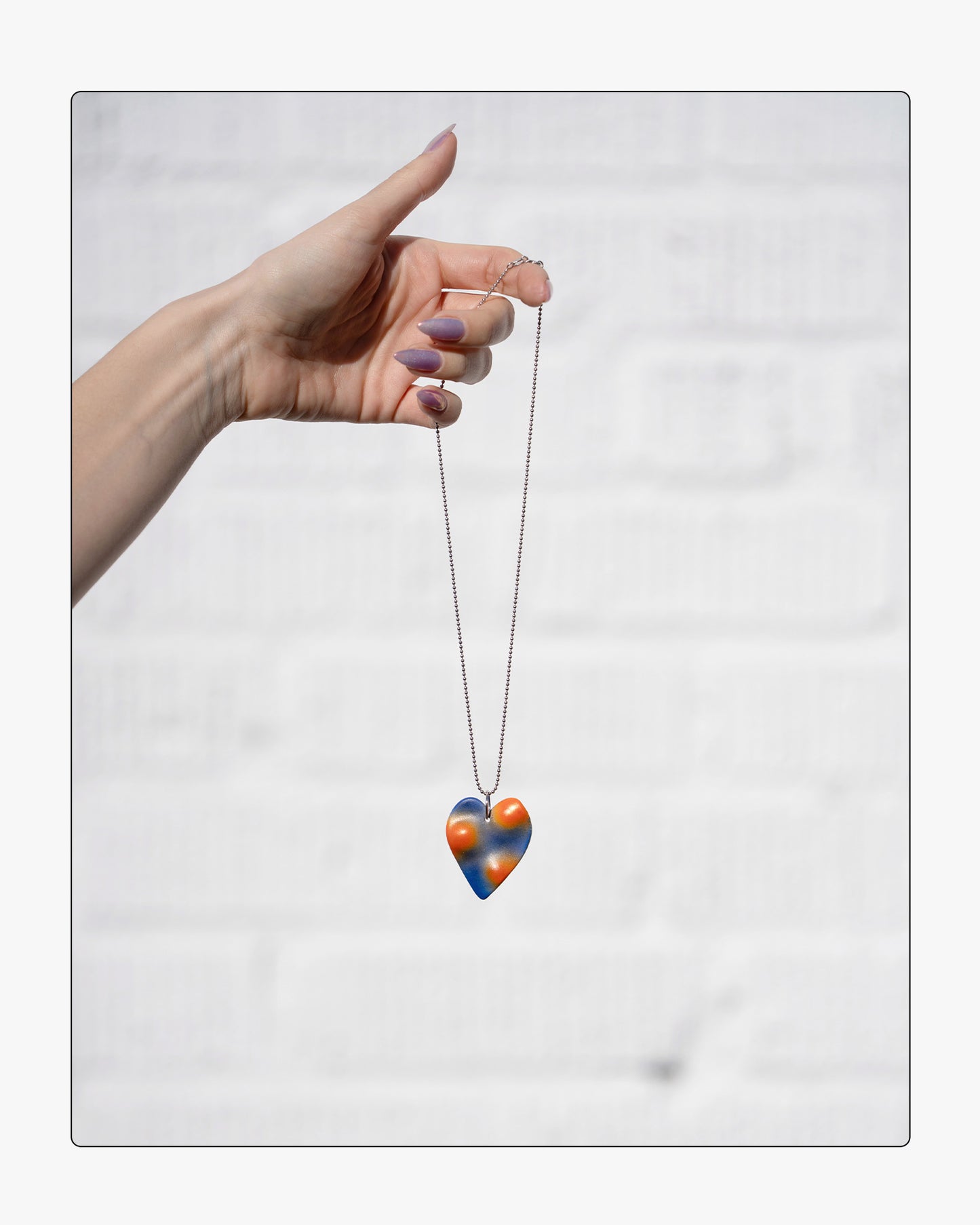 Hand holding a the Topo heart necklace, showing the 18" silver ball chain against a white background.